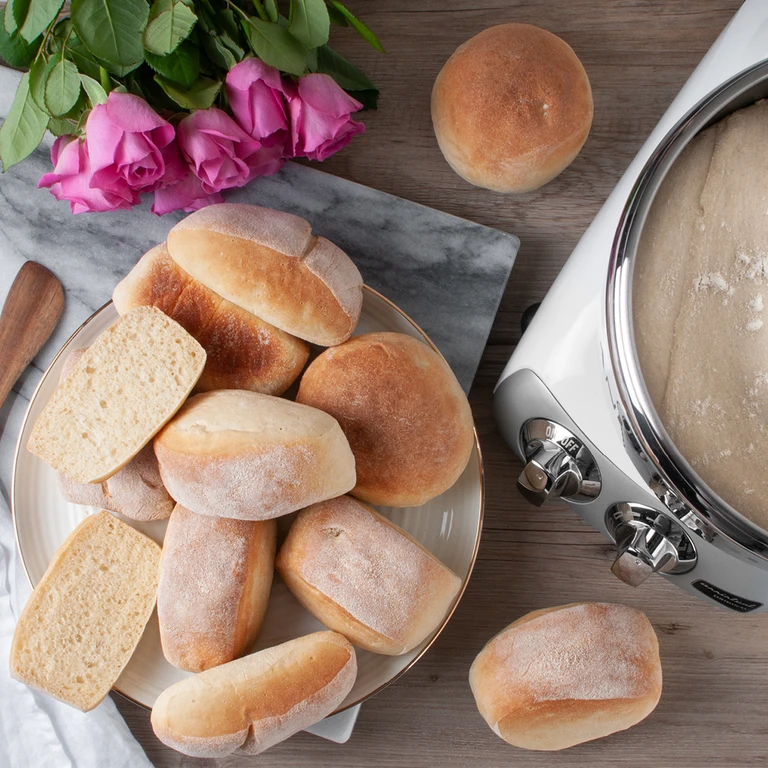 Freshly baked bread rolls on a plate beside an Ankarsrum stand mixer bowl with dough, pink roses in background.