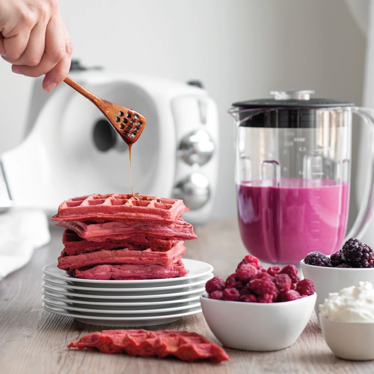 Stacked pink berry waffles drizzled with honey, beside a blender with berry smoothie, raspberries, blackberries, and cream.