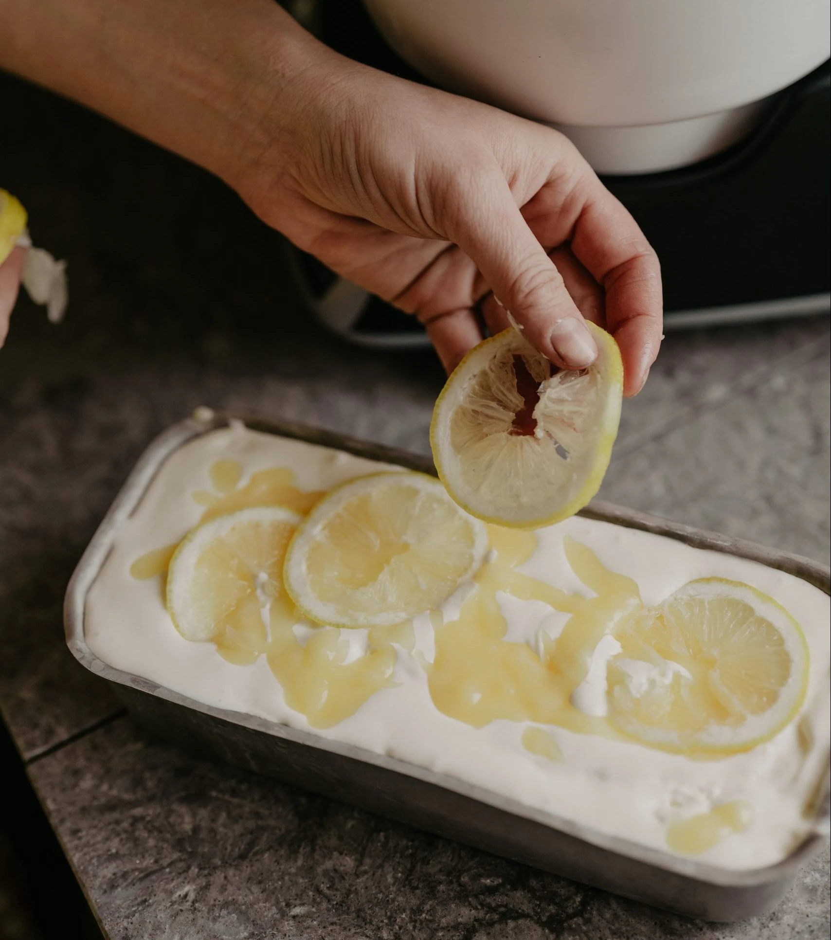 Hand placing lemon slices on creamy dessert in loaf pan with lemon curd, prepared using Ankarsrum stand mixer.