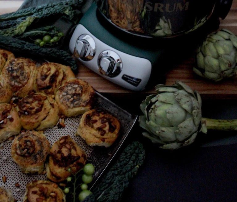 Ankarsrum stand mixer surrounded by savory pinwheel rolls on a baking tray, fresh artichokes, and dark leafy kale greens.