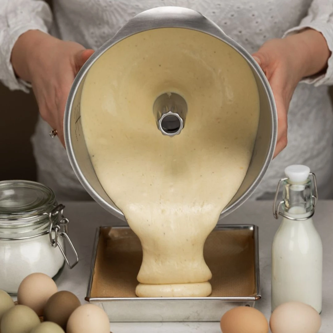 Luftig kakerøre helles fra en miksebolle ned i en firkantet bakeform. Rundt formen ligger egg, melk og sukker klare til baking.