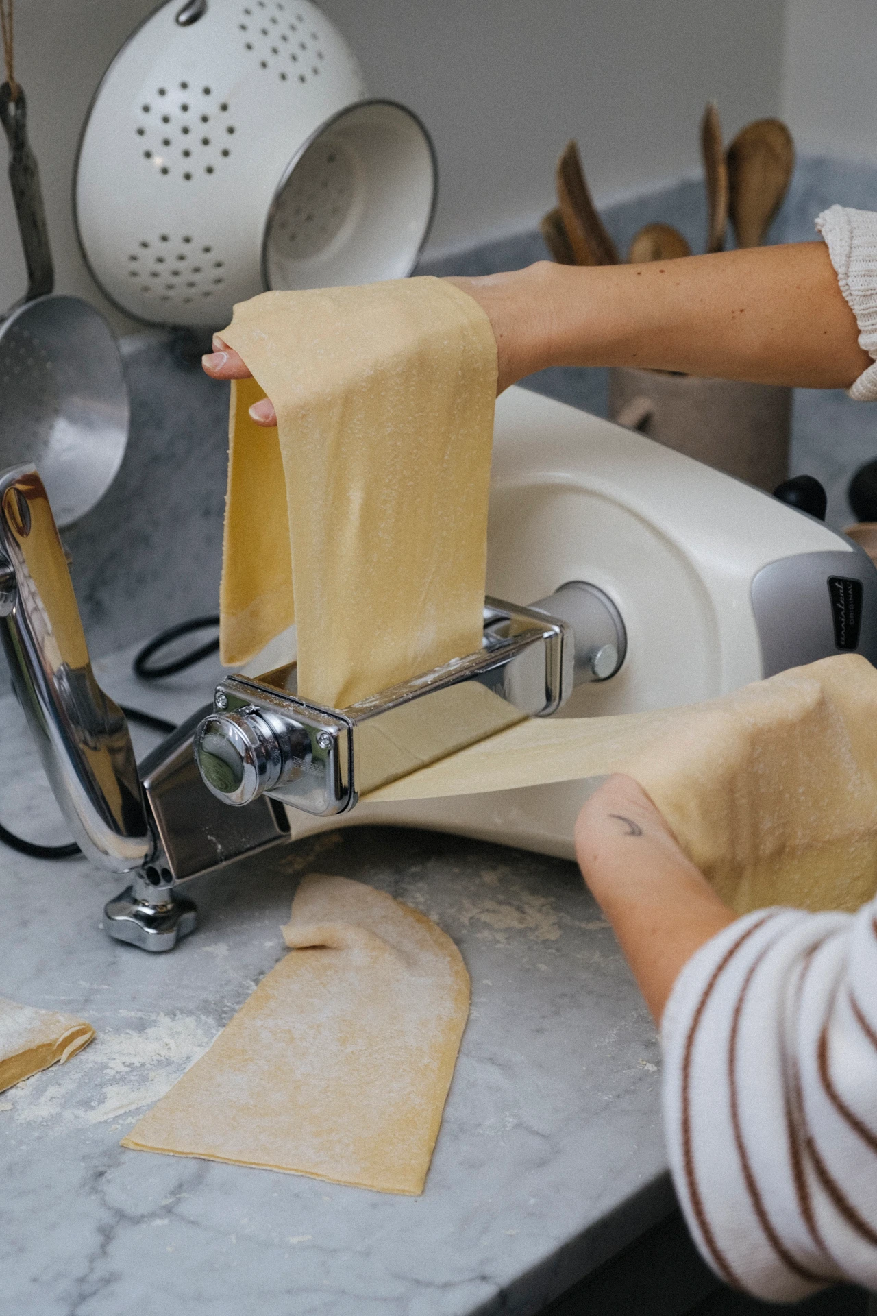 Hands feeding fresh pasta dough through an Ankarsrum stand mixer pasta roller attachment on a marble countertop.