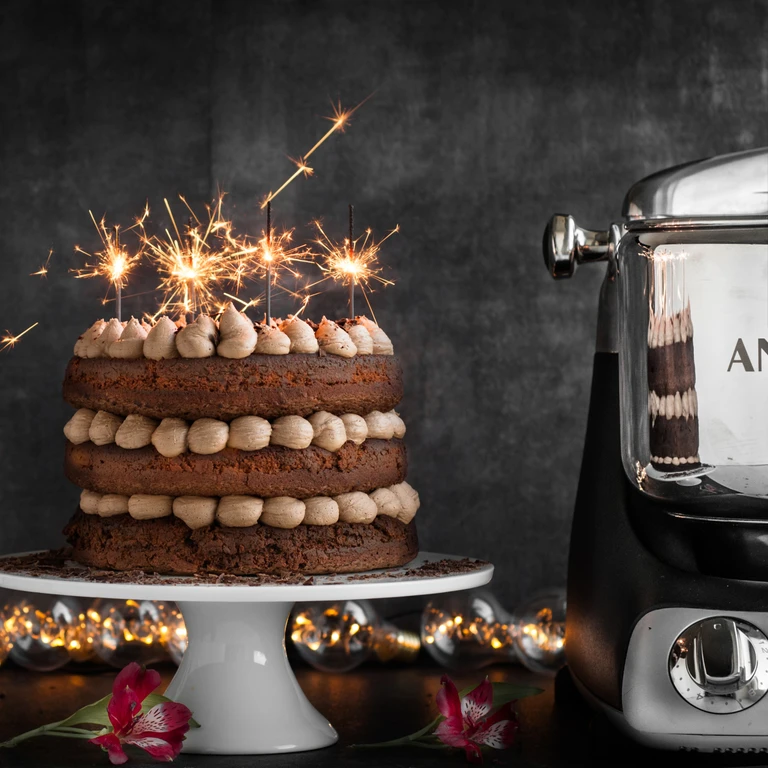 Chocolate layer cake with sparklers on a white stand beside an Ankarsrum stand mixer, surrounded by silver ornaments.