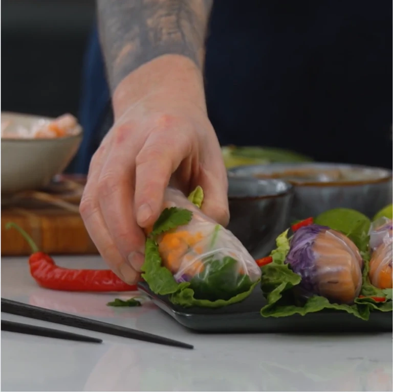 Tattooed chef placing fresh Vietnamese spring rolls with shrimp, herbs, and vegetables on a dark serving tray.