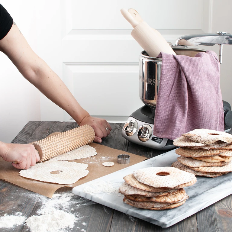 Person rolling Swedish crispbread dough with a textured rolling pin beside an Ankarsrum Original stand mixer.