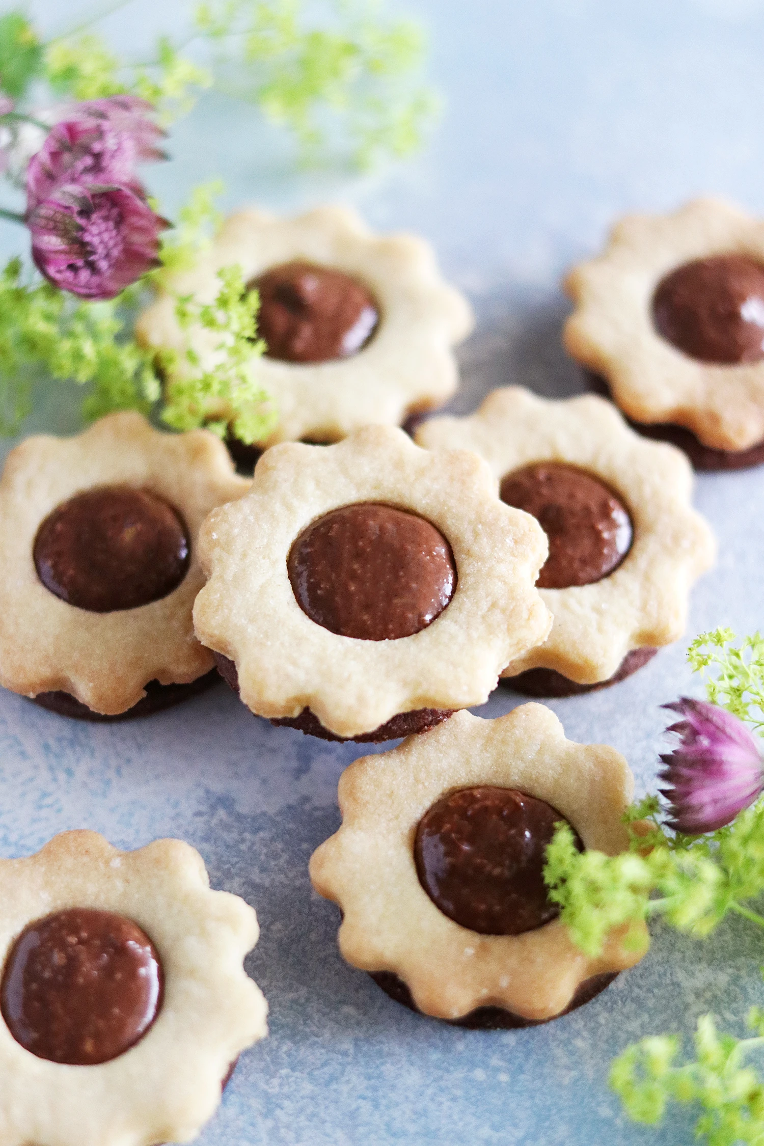 Flower-shaped shortbread cookies with chocolate ganache centers, arranged on a blue surface with purple wildflowers.