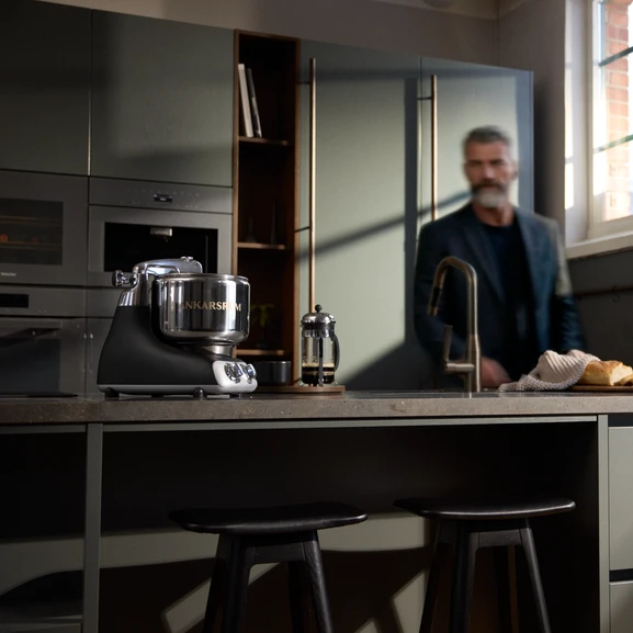 Ankarsrum Assistent Original en noir sur un îlot de cuisine dans une cuisine sombre, avec un homme visible à l’arrière-plan.