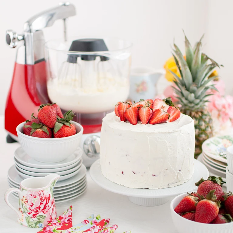 Ankarsrum red stand mixer with whisk attachment beside a white frosted strawberry cake on a cake stand.