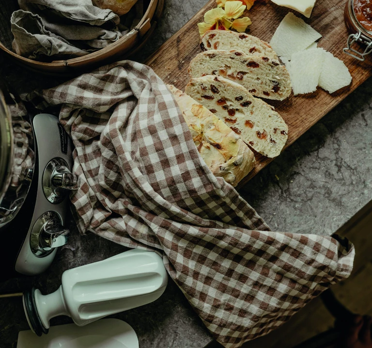 Sliced fruit bread on wooden board with cheese, jam jar, and Ankarsrum stand mixer beside gingham kitchen towel.