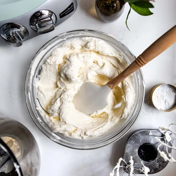 Fluffy white frosting in a glass bowl with a spatula, beside an Ankarsrum Original stand mixer on a marble surface.