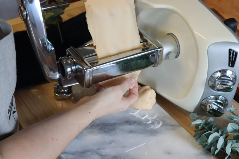 Hands feeding pasta dough through an Ankarsrum pasta roller attachment on a cream-colored stand mixer.