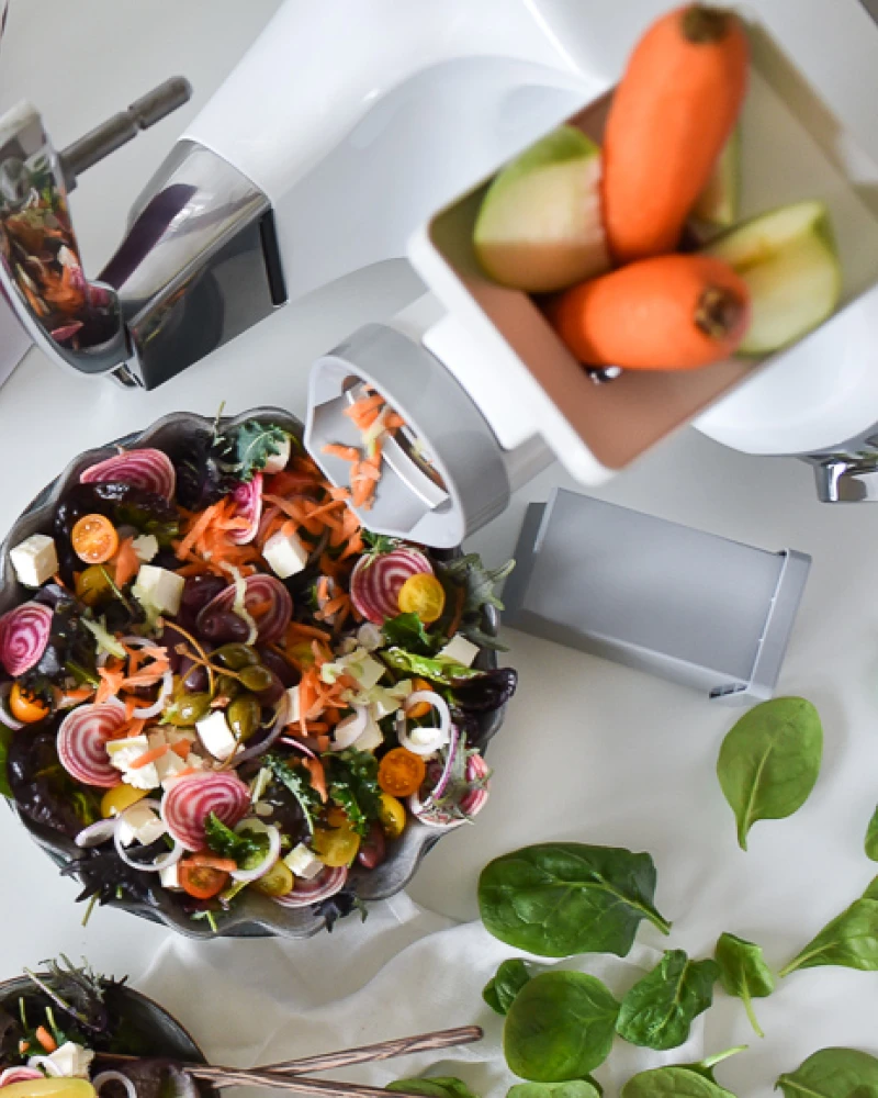 Ankarsrum stand mixer with salad prep attachment shredding carrots into a colorful bowl of fresh vegetables and feta.