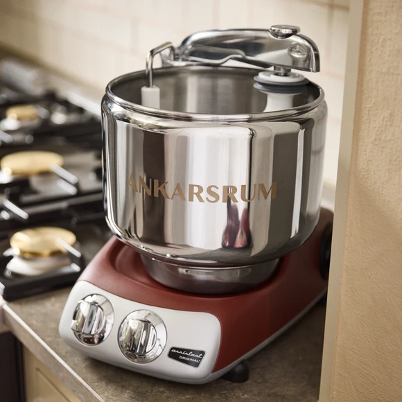 Ankarsrum Assistant Original stand mixer in red with stainless steel bowl, displayed on a kitchen countertop near a gas stove.