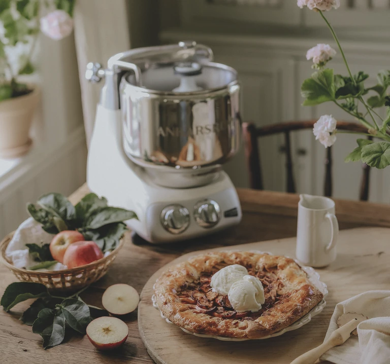 Ankarsrum stand mixer beside a rustic apple galette topped with ice cream, fresh apples, and pink flowers on a wooden table.