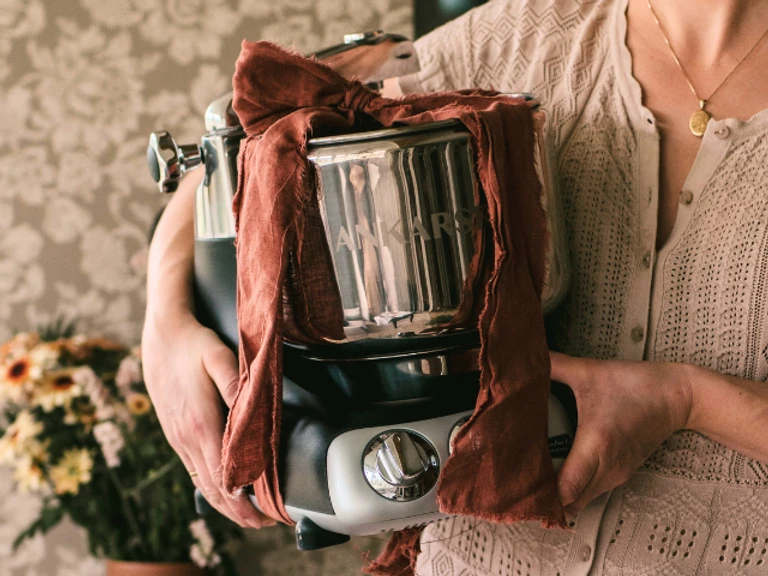 Woman holding an Ankarsrum stand mixer wrapped in a rustic red cloth, showcasing the iconic kitchen appliance.