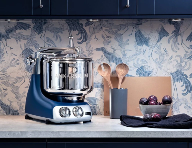 Ankarsrum stand mixer in navy blue and chrome on a kitchen counter with wooden spoons, plums, and crane-patterned wallpaper.