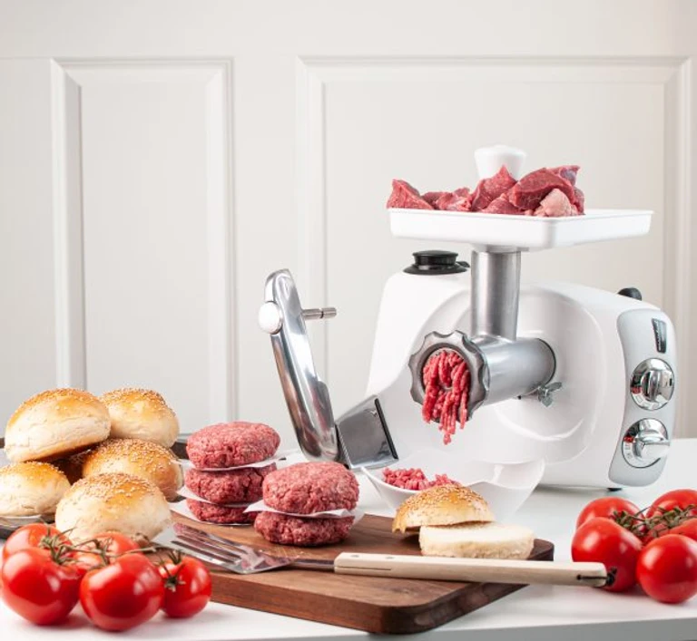 Ankarsrum stand mixer with meat grinder attachment, grinding fresh beef into burger patties, with buns and tomatoes nearby.