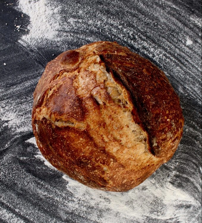 Freshly baked rustic sourdough boule with golden crust on flour-dusted dark surface, made with Ankarsrum stand mixer.