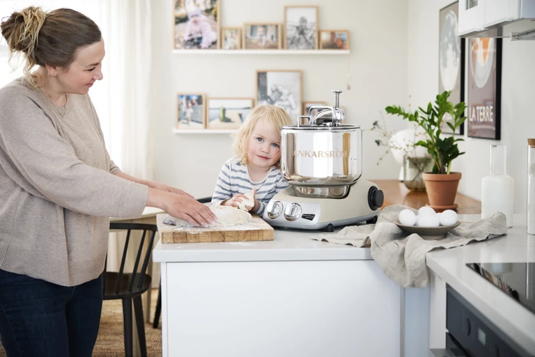 Mother and toddler baking bread dough together using an Ankarsrum stand mixer on a white kitchen counter.
