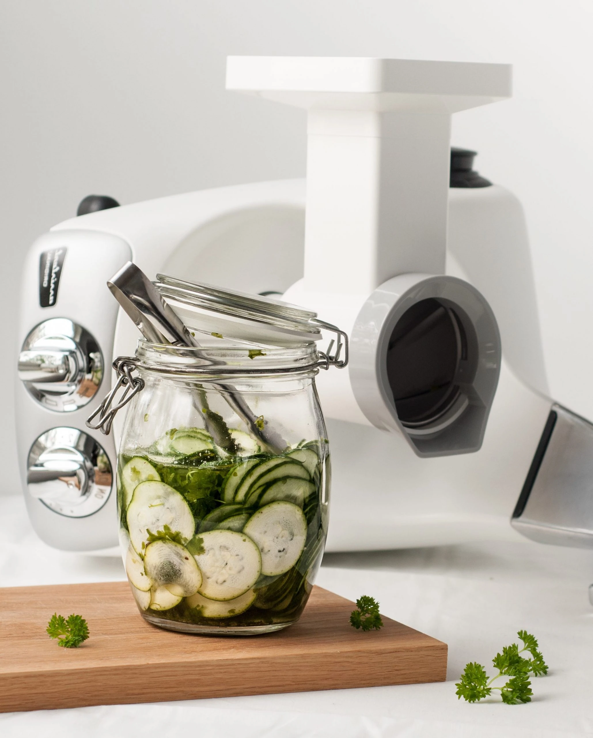 Pickled cucumber slices in a glass jar on a wooden board beside a white Ankarsrum stand mixer with slicer/shredder attachment.
