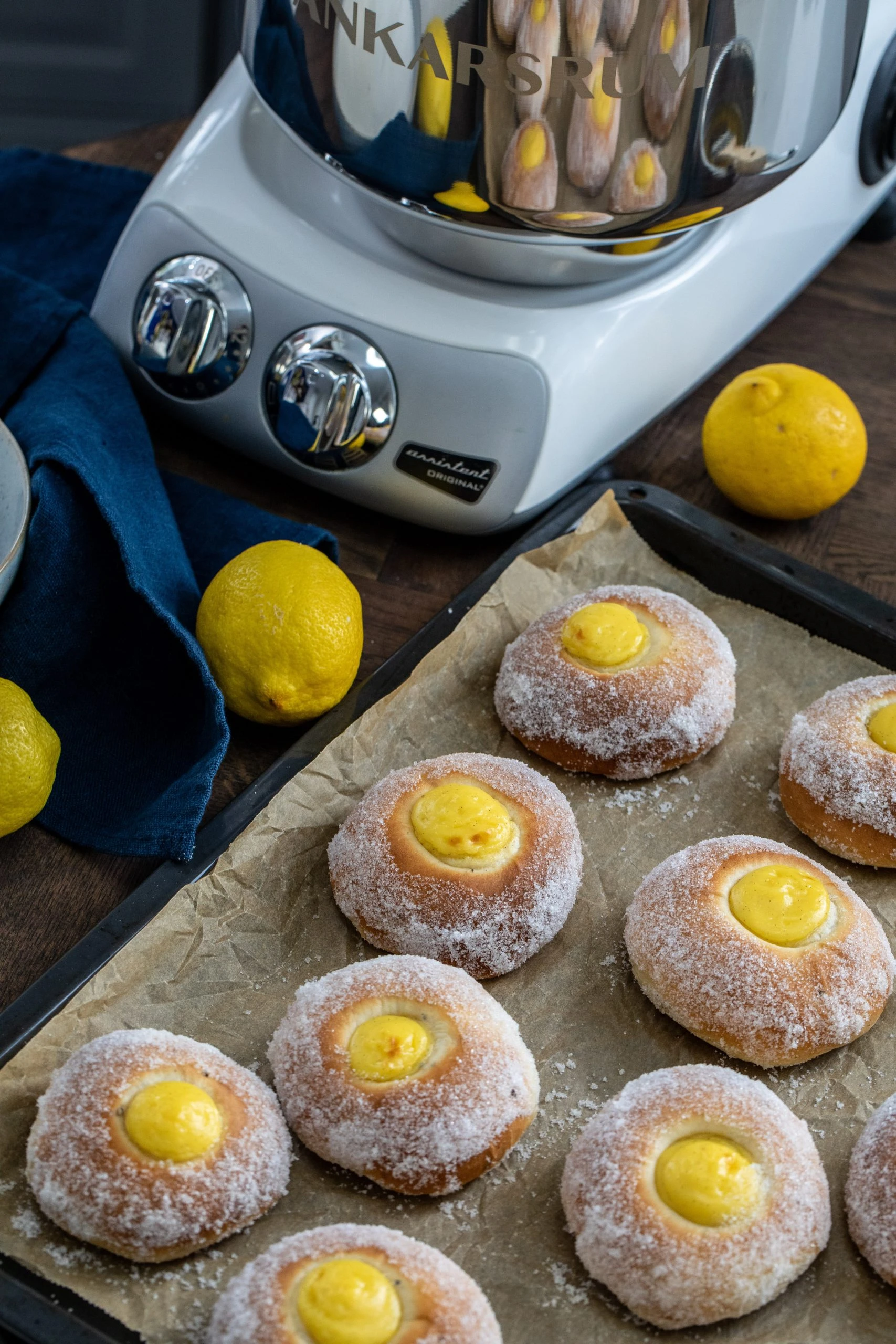 Ankarsrum stand mixer beside a tray of sugar-coated lemon curd donuts on parchment paper, with fresh lemons nearby.