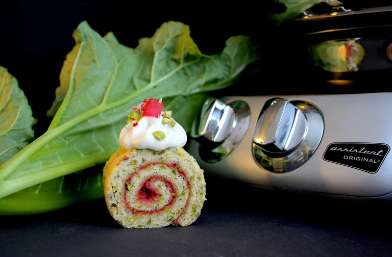 Ankarsrum Assistent Original stand mixer beside a pistachio rhubarb Swiss roll slice topped with cream, rhubarb, and pistachios.