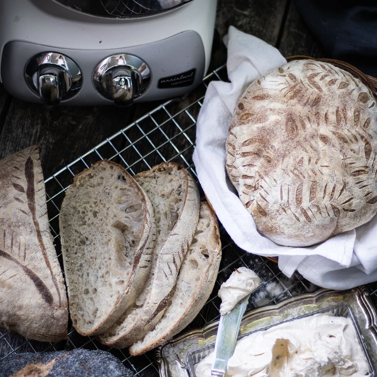 Freshly baked artisan sourdough bread with decorative scoring, sliced open, beside an Ankarsrum Original stand mixer.