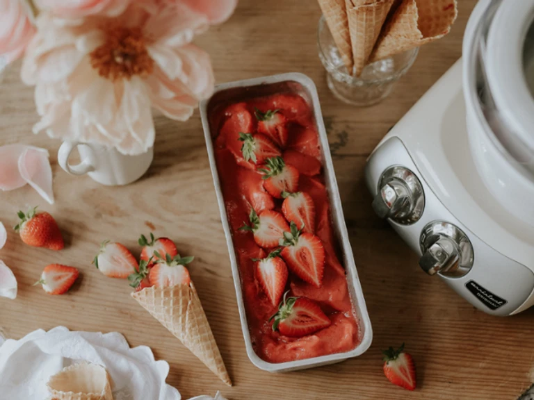 Homemade strawberry sorbet in a loaf pan with fresh strawberries, waffle cones, and an Ankarsrum machine on a wooden surface.