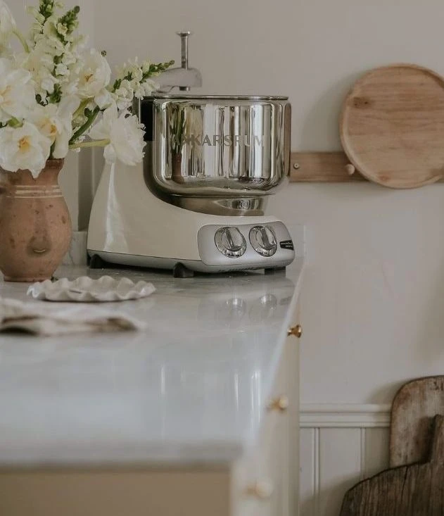 Ankarsrum stand mixer in creme with chrome bowl on marble countertop, beside white flowers in a terracotta vase.