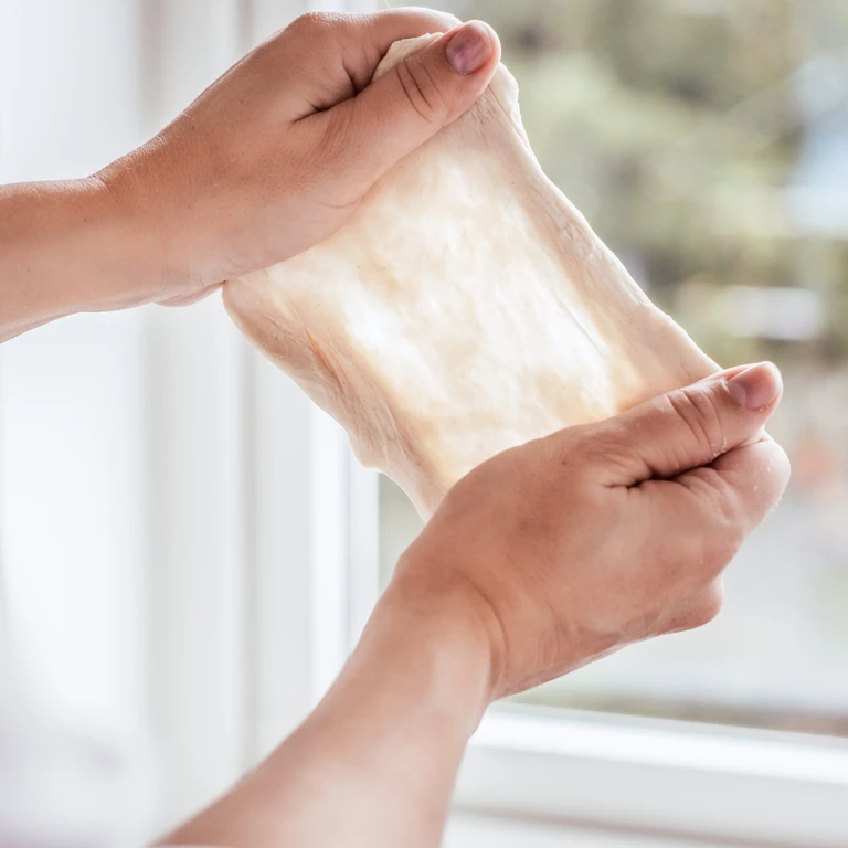 Hands performing the windowpane test on smooth, elastic bread dough, demonstrating perfect gluten development with an Ankarsrum mixer.
