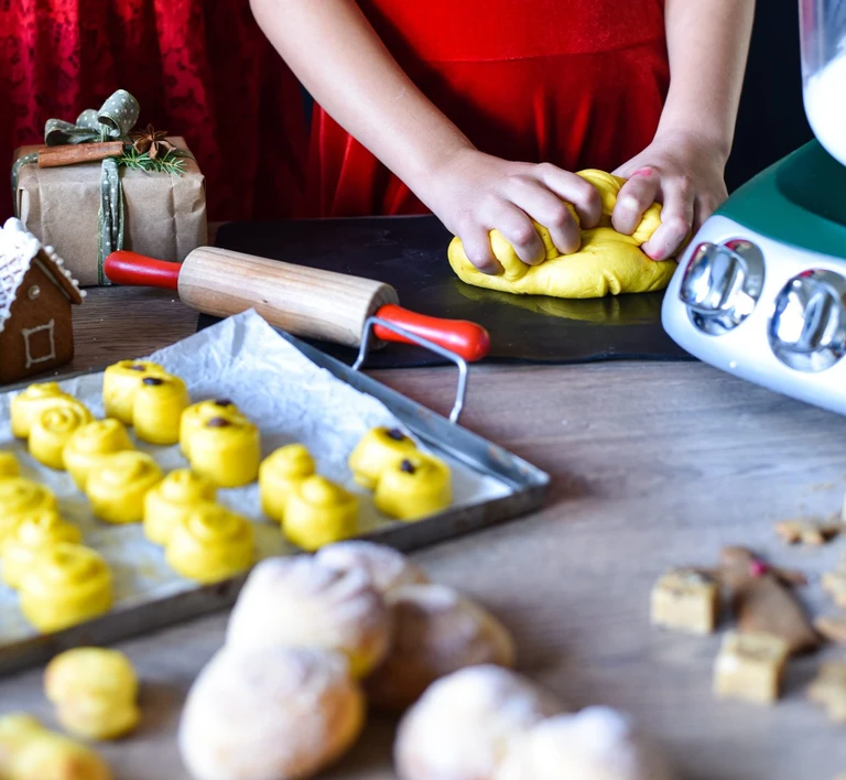 Hands kneading yellow saffron dough beside a tray of unbaked St. Lucia buns, rolling pin, and Ankarsrum stand mixer.