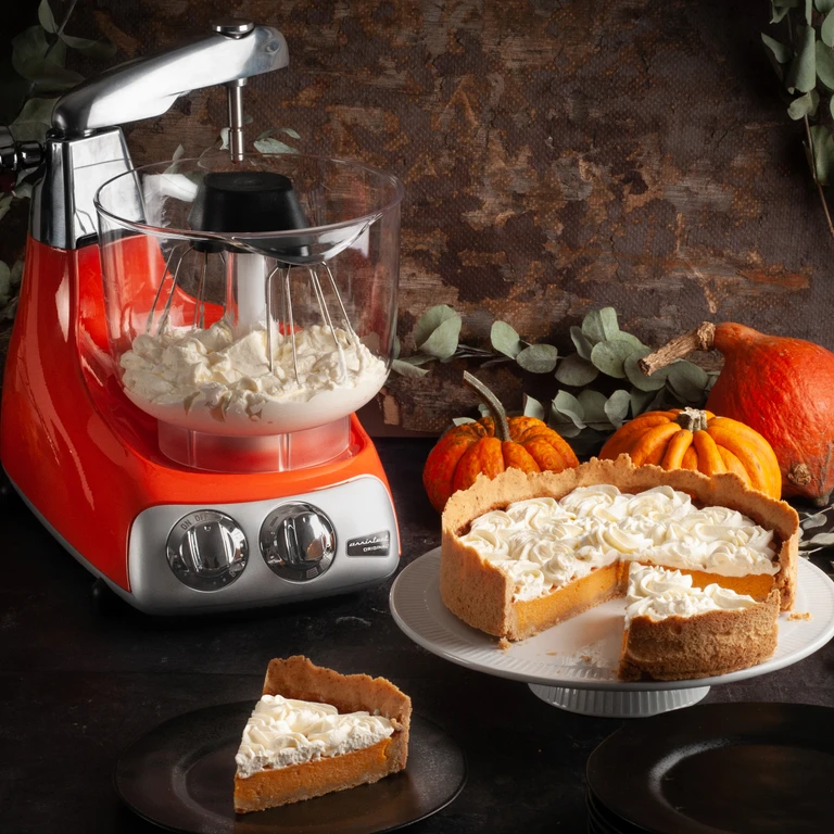 Ankarsrum Original red stand mixer whipping cream beside a pumpkin pie topped with whipped cream rosettes.