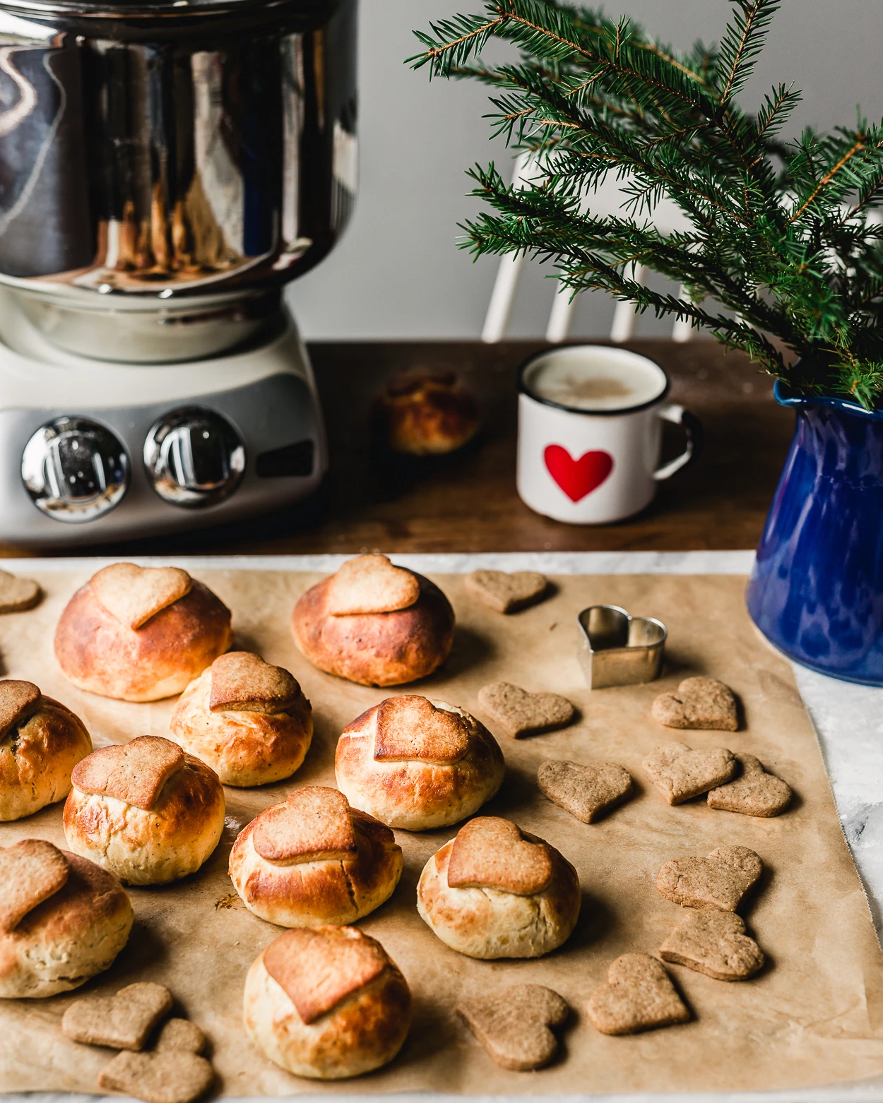 Freshly baked heart-shaped bread rolls and gingerbread cookies on parchment paper beside an Ankarsrum stand mixer.
