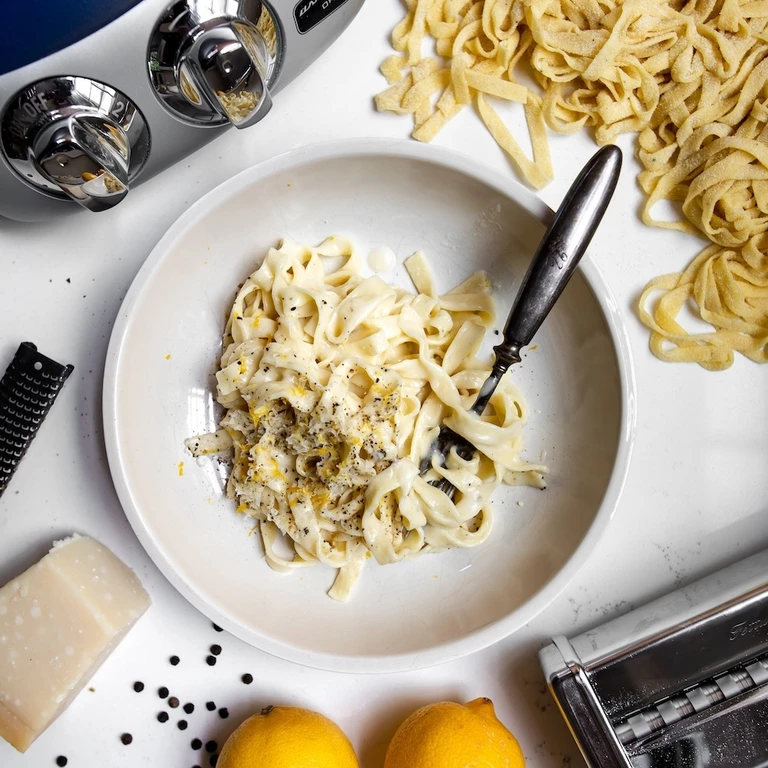 Lemon pepper fettuccine pasta in a white bowl with Ankarsrum pasta roller, grater, lemons, and Parmesan cheese nearby.