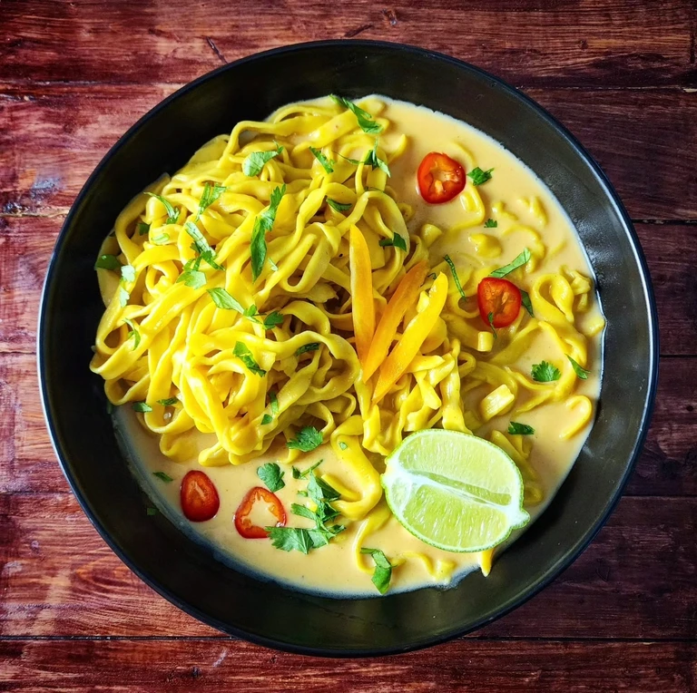 Golden turmeric noodles in creamy coconut broth, garnished with chili, lime, and cilantro, made with Ankarsrum pasta attachment.