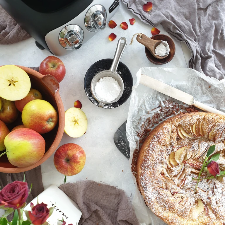 Freshly baked apple cake dusted with powdered sugar beside a bowl of apples, roses, and an Ankarsrum kitchen appliance.