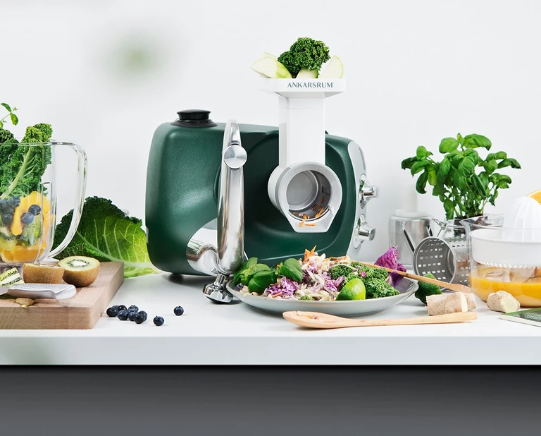 Ankarsrum forest green stand mixer with vegetable grater attachment, surrounded by fresh vegetables, fruits, a colorful salad bowl, citrus juicer and blender attachments.