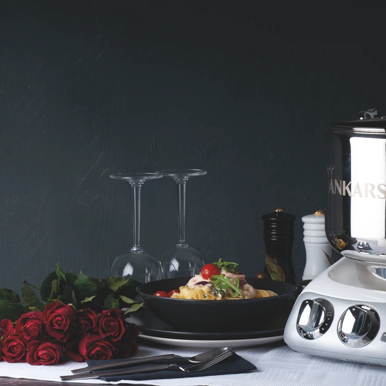 Ankarsrum stand mixer beside a romantic pasta dish, red roses, wine glasses, and pepper mills on an elegant dark table.