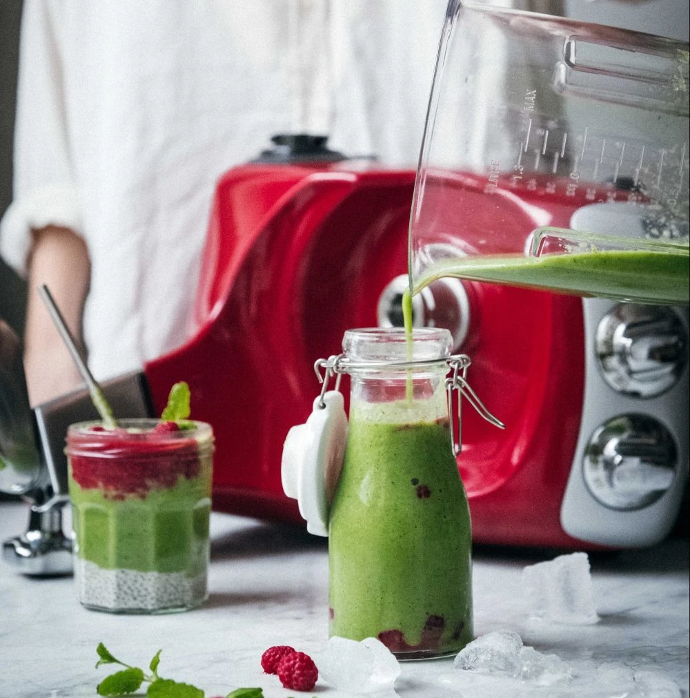 Ankarsrum red blender pouring green smoothie into glass jar with raspberries and chia seeds on marble surface.