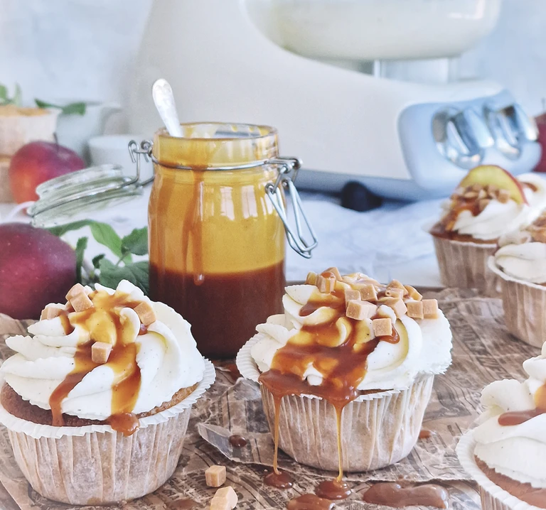 Caramel drizzled cupcakes with white frosting beside a jar of caramel sauce, apples, and an Ankarsrum stand mixer in background.