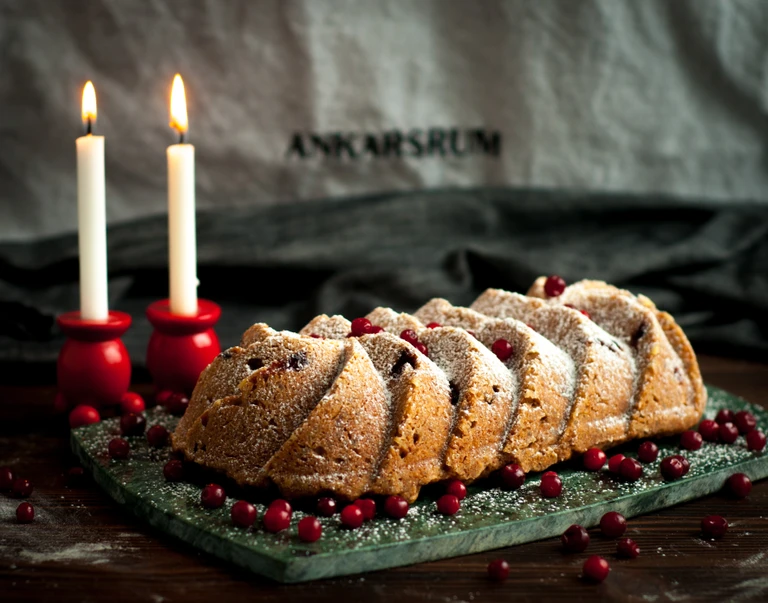 Sliced cranberry bundt cake dusted with powdered sugar on marble board with lit candles; Ankarsrum branded linen backdrop.
