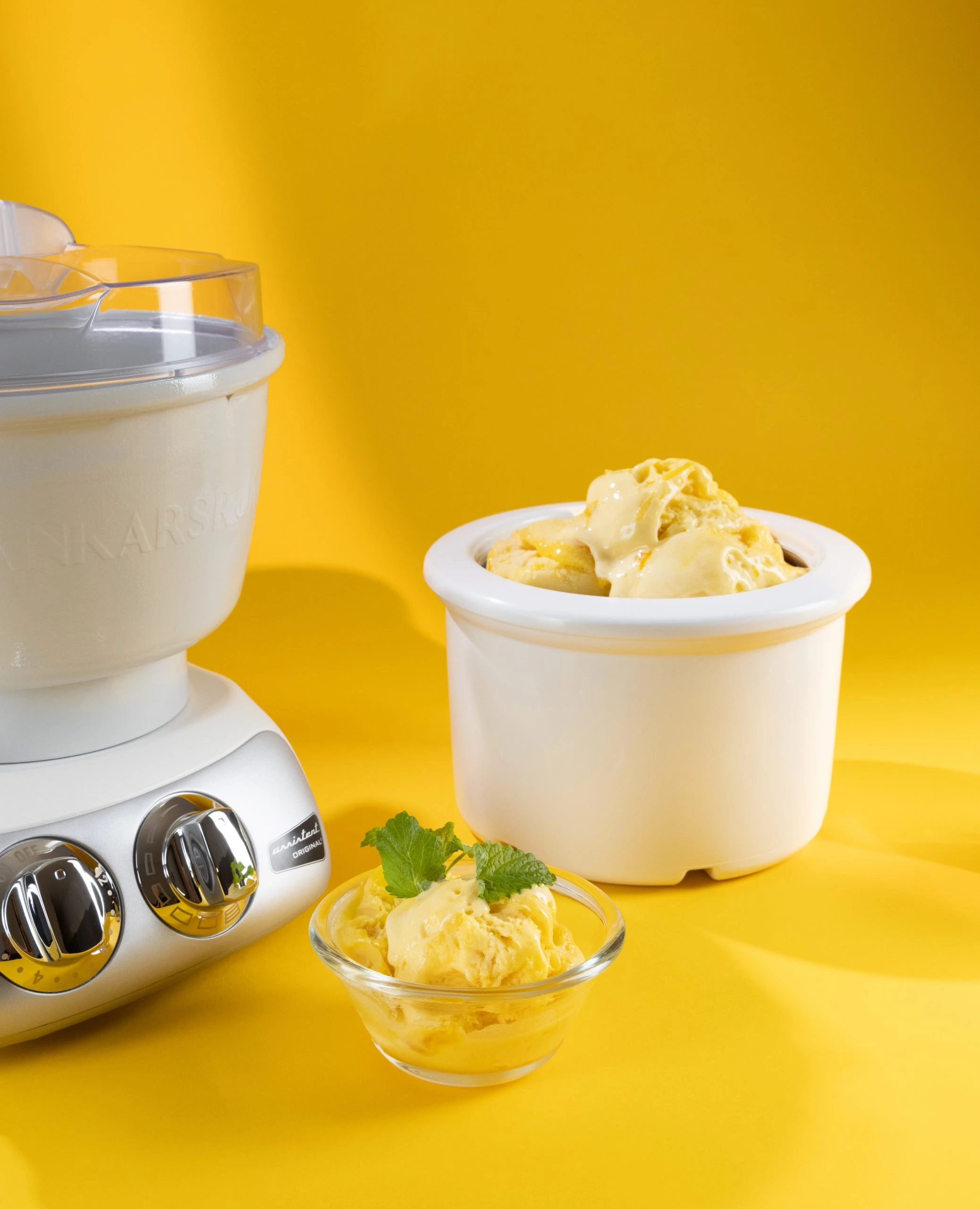 Ankarsrum Original stand mixer with ice cream maker attachment beside a bowl of fresh yellow mango ice cream garnished with mint.