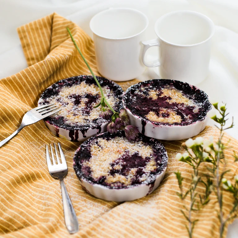 Three individual blueberry crumble desserts in white ramekins on a golden cloth, made with Ankarsrum stand mixer.