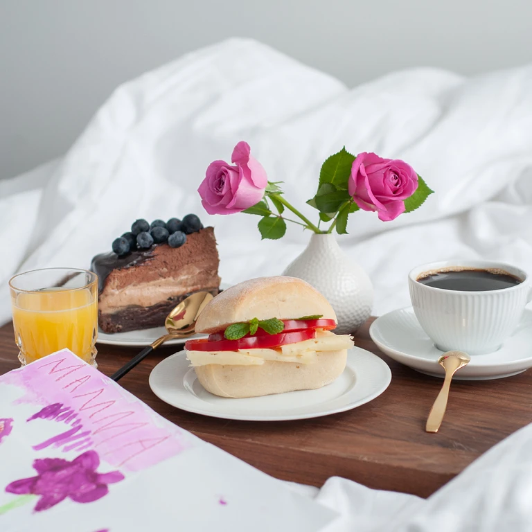 Breakfast in bed tray with sandwich, chocolate cake, orange juice, coffee, pink roses, and a handmade Mama card.