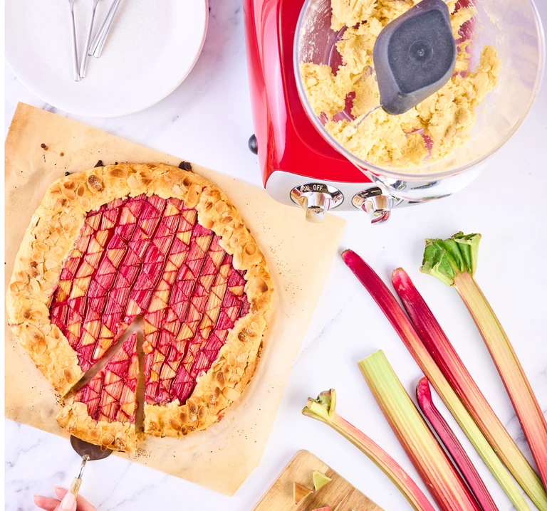 Rhubarb galette being sliced beside fresh rhubarb stalks and a red Ankarsrum stand mixer with dough.