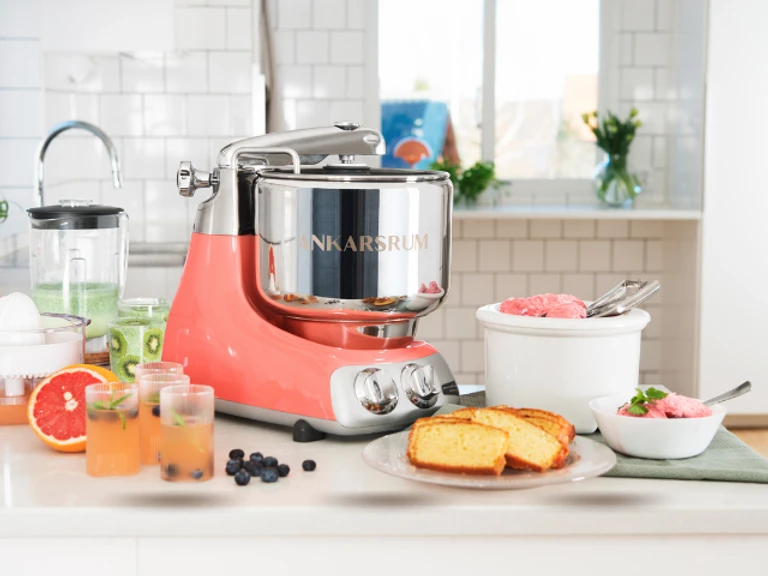 Ankarsrum stand mixer in coral crush with stainless steel bowl, surrounded by fresh fruits, bread, and ice cream on a kitchen counter.