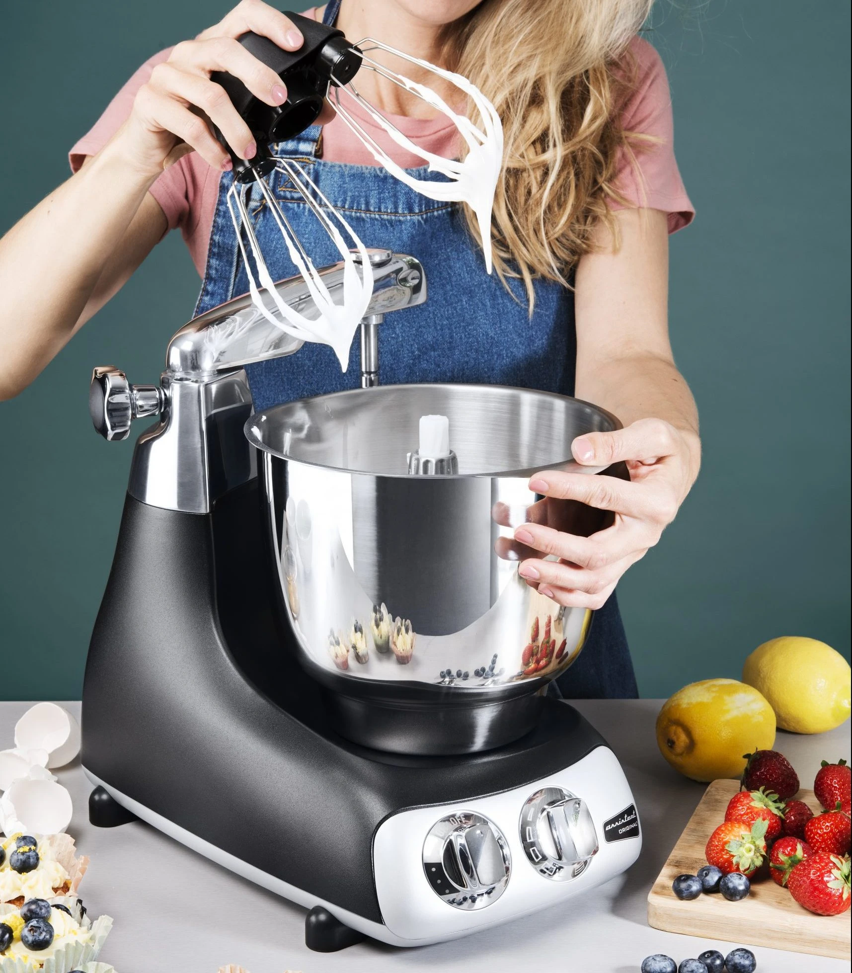 Woman removing balloon whisk attachment from Ankarsrum Original stand mixer, with whipped cream, strawberries, blueberries, and lemons nearby.