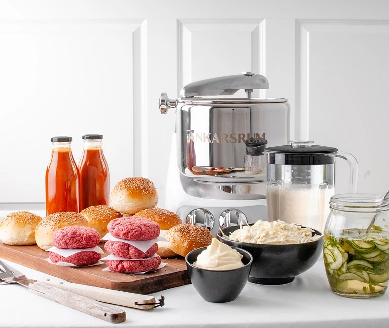 Ankarsrum stand mixer surrounded by homemade burger buns, raw patties, sauces, coleslaw, mayo, and pickled cucumbers.
