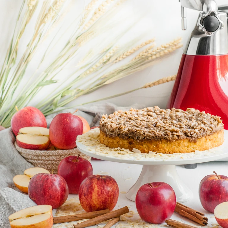 Apple crumble cake on white stand, surrounded by red apples and cinnamon sticks, with red Ankarsrum stand mixer in background.