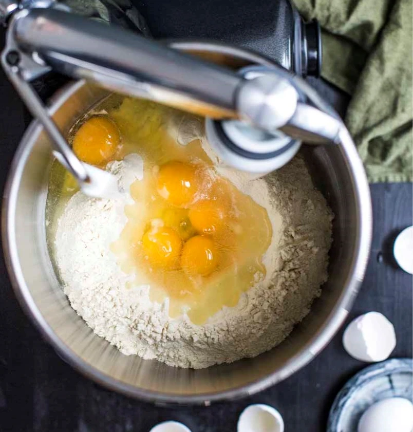 Ankarsrum stand mixer bowl with cracked eggs and flour, ready for dough mixing, with eggshells beside it.